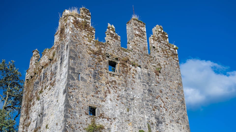 A tall, weathered stone castle with battlements and a cross on top, set against a clear blue sky.