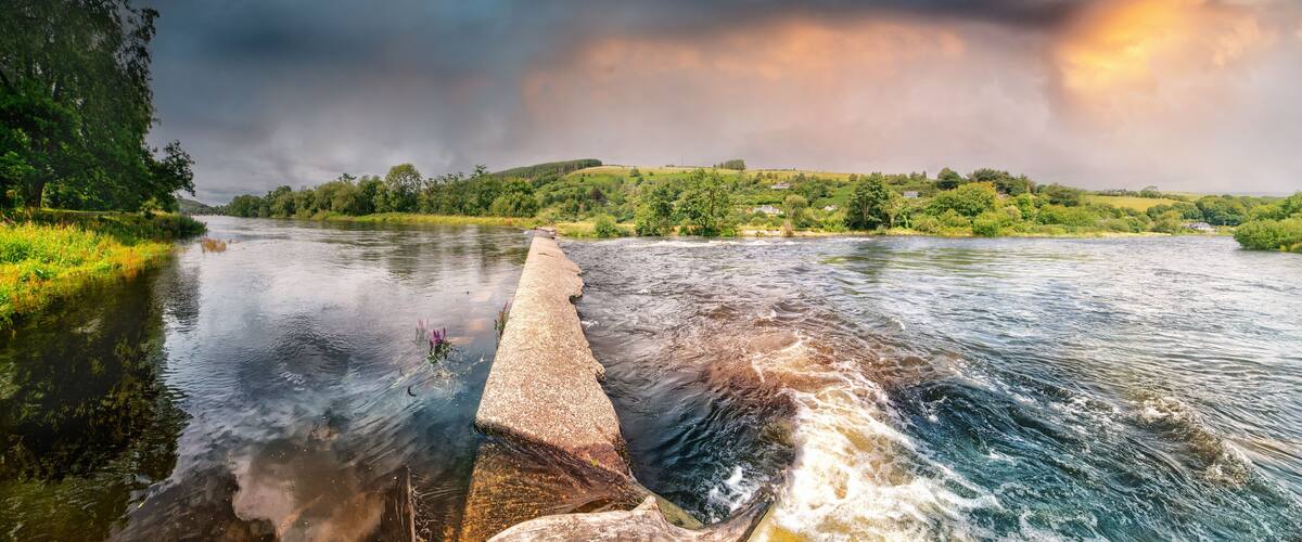 A river with a concrete weir, surrounded by lush greenery and a cloudy sky.