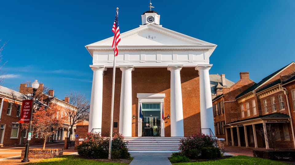 A white neoclassical building with columns and a clock tower, surrounded by brick buildings and a clear blue sky.