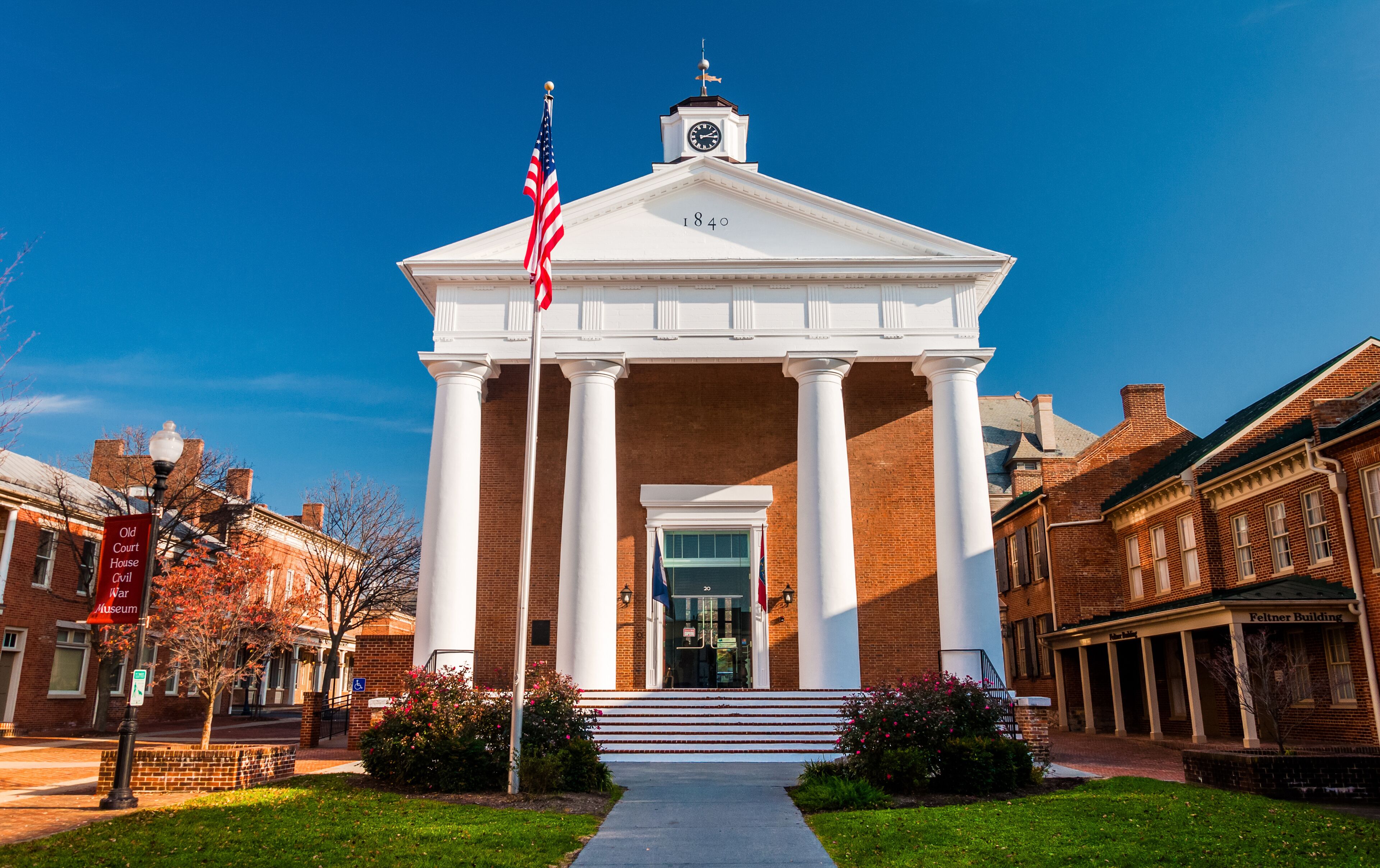 A white neoclassical building with columns and a clock tower, surrounded by brick buildings and a clear blue sky.