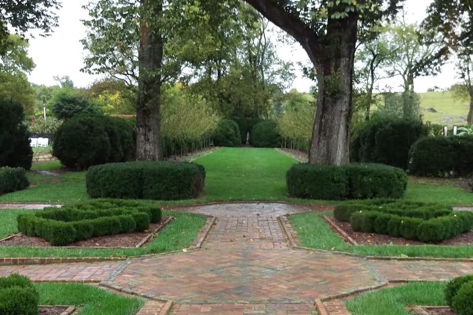 A brick pathway leading to a central lawn area, surrounded by trimmed hedges and trees.