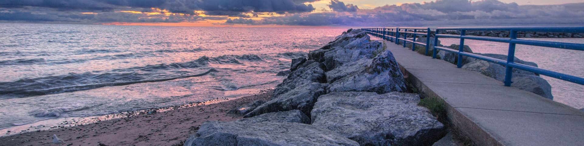 A rocky shoreline with a blue railing, a sunset over the ocean, and a cloudy sky.