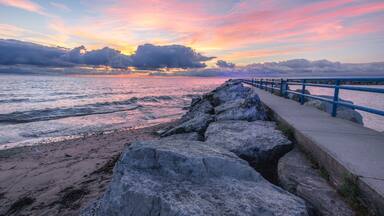 A rocky shoreline with a blue railing, a sunset over the ocean, and a cloudy sky.