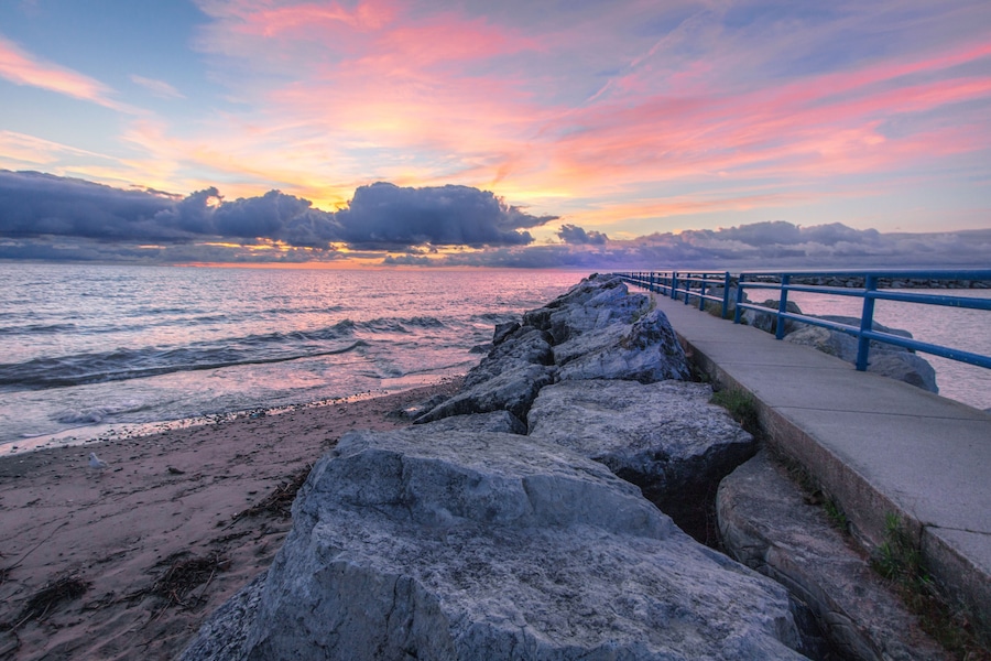 A rocky shoreline with a blue railing, a sunset over the ocean, and a cloudy sky.