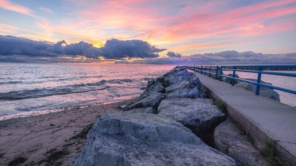 A rocky shoreline with a blue railing, a sunset over the ocean, and a cloudy sky.