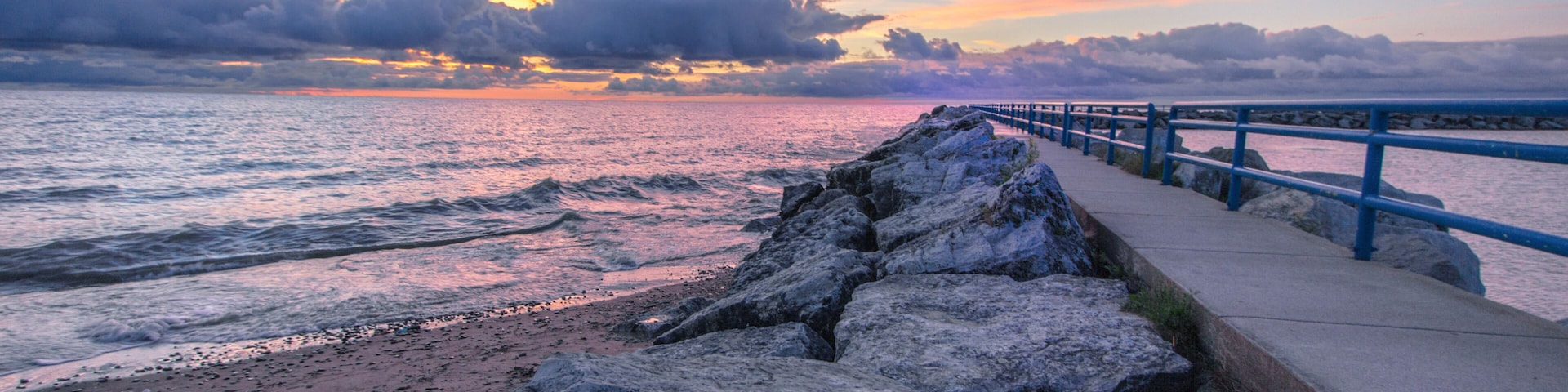 A rocky shoreline with a blue railing, a sunset over the ocean, and a cloudy sky.