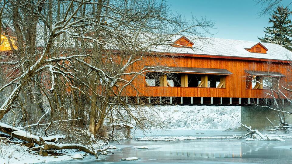 A wooden covered bridge over a frozen river, surrounded by snow-covered trees.