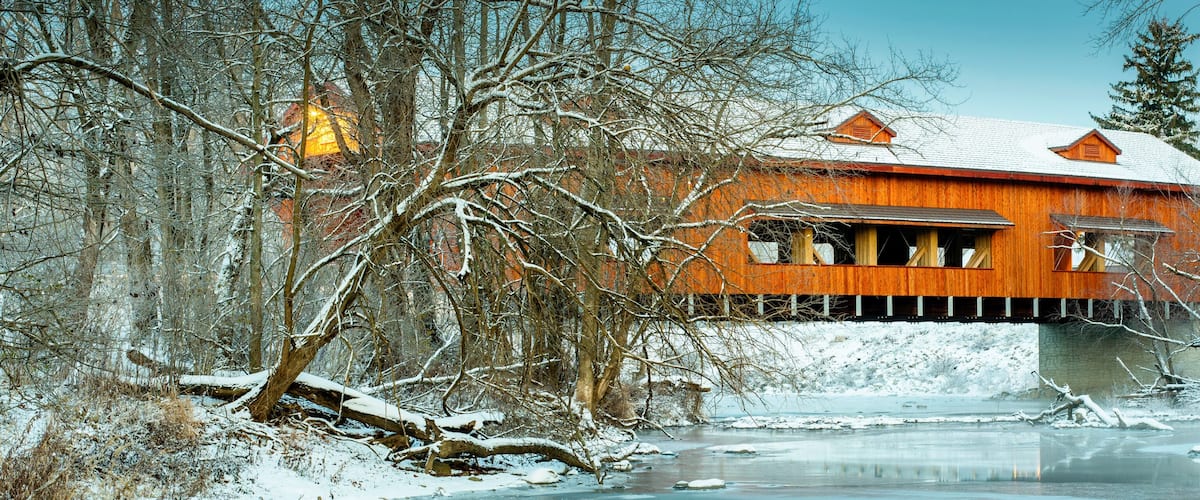 A wooden covered bridge over a frozen river, surrounded by snow-covered trees.