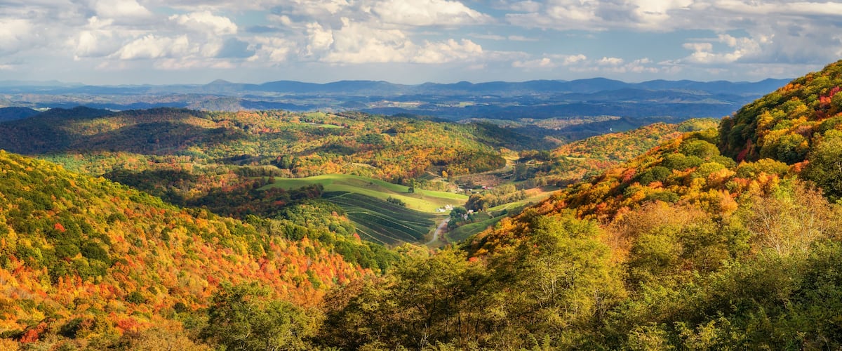 A valley with autumn foliage, rolling hills, and distant mountains under a partly cloudy sky.