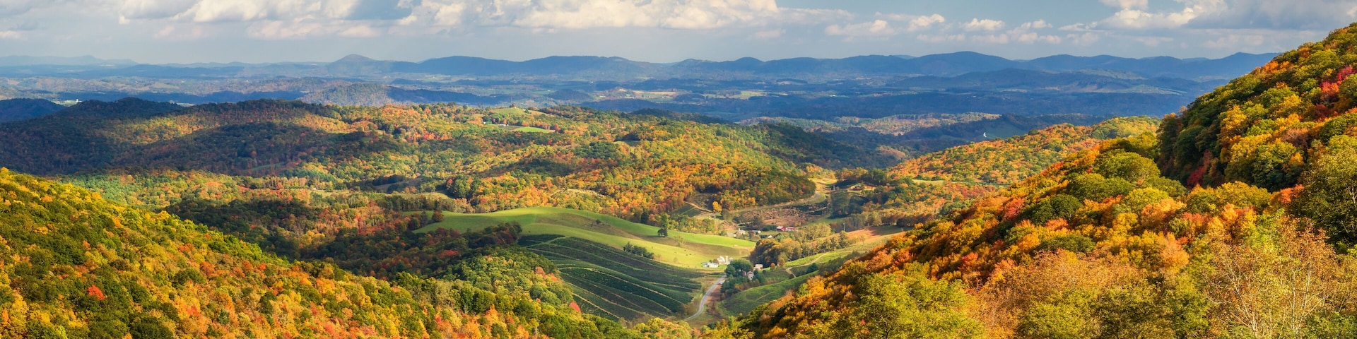 A valley with autumn foliage, rolling hills, and distant mountains under a partly cloudy sky.