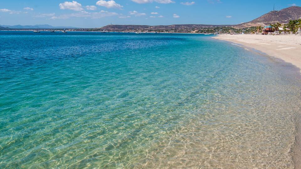 A clear turquoise sea, a sandy beach, and a distant mountain range.