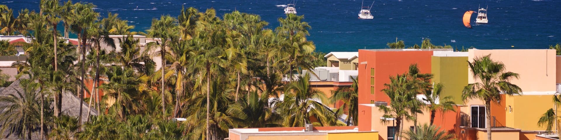 A coastal town with palm trees, colorful buildings, and a view of the ocean with kitesurfers.