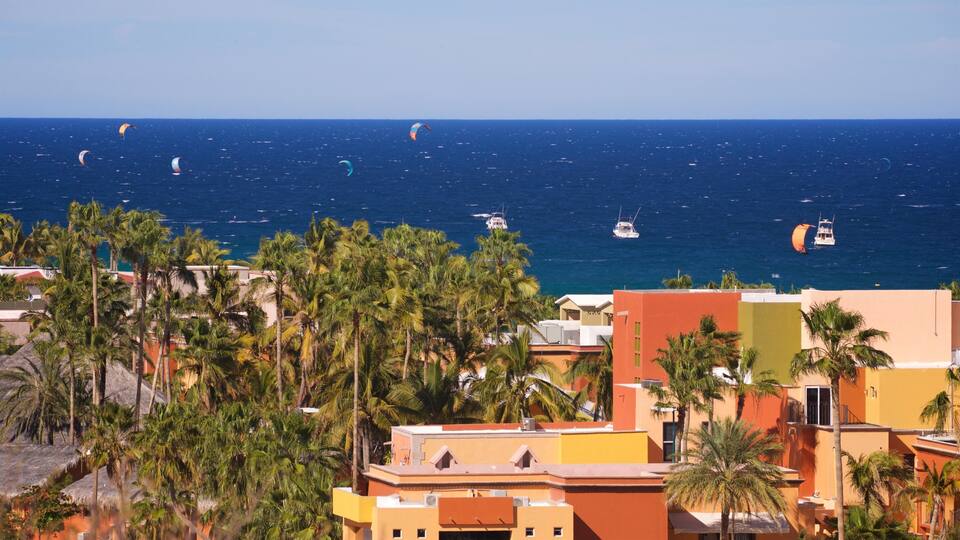 A coastal town with palm trees, colorful buildings, and a view of the ocean with kitesurfers.