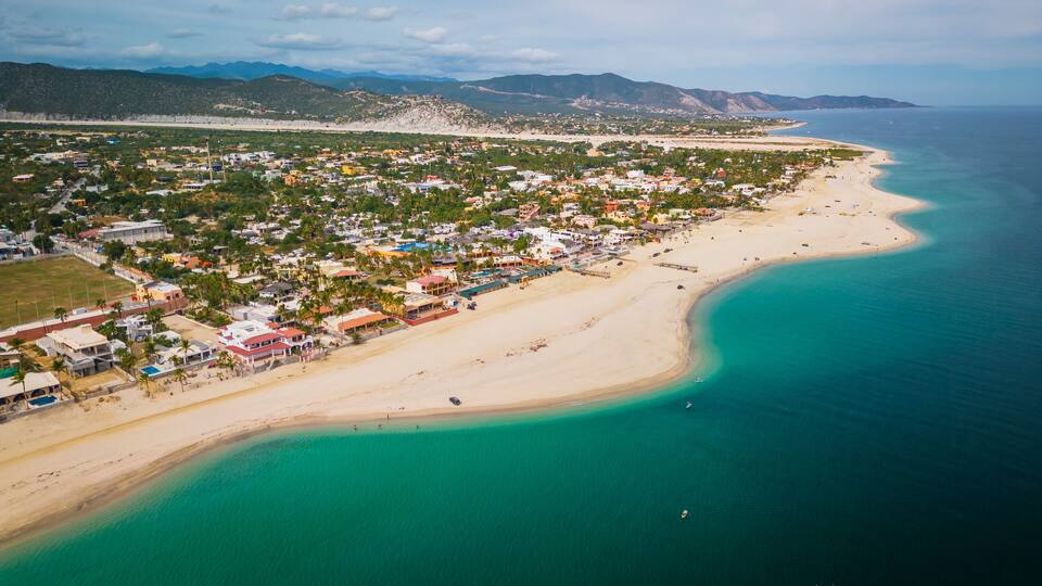 An aerial view of a coastal town with a beach, houses, and mountains in the background.