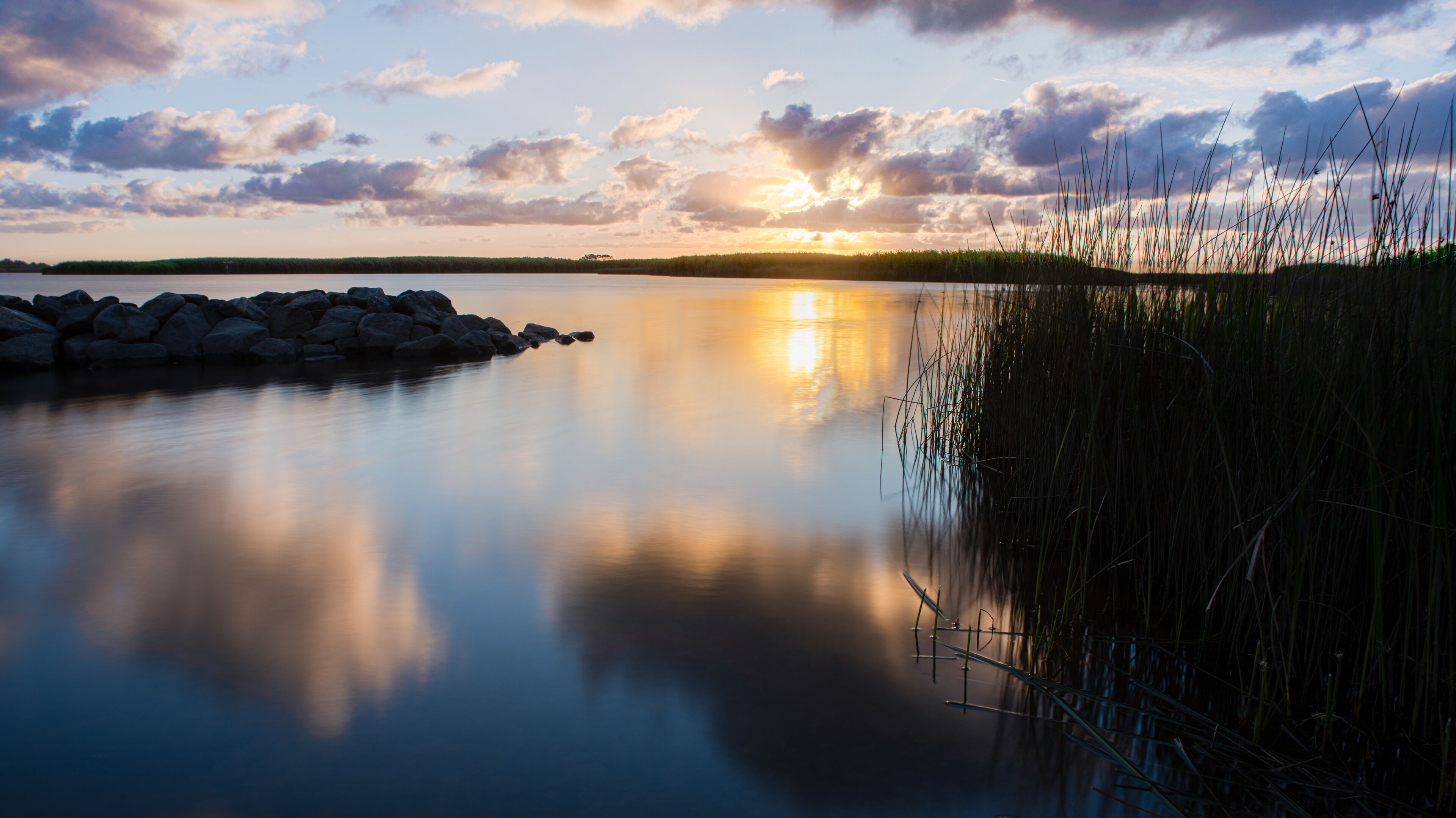 A calm lake at sunset with a rocky shoreline and tall grasses reflecting the sky.