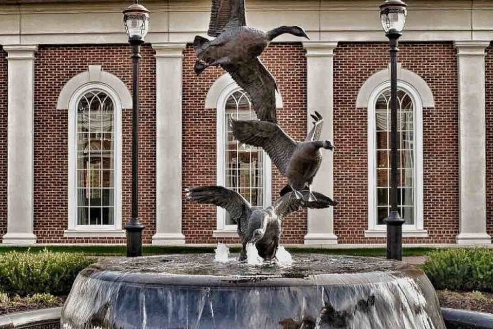 Una fuente con una escultura de pájaro frente a un edificio de ladrillo con ventanas arqueadas y faroles.