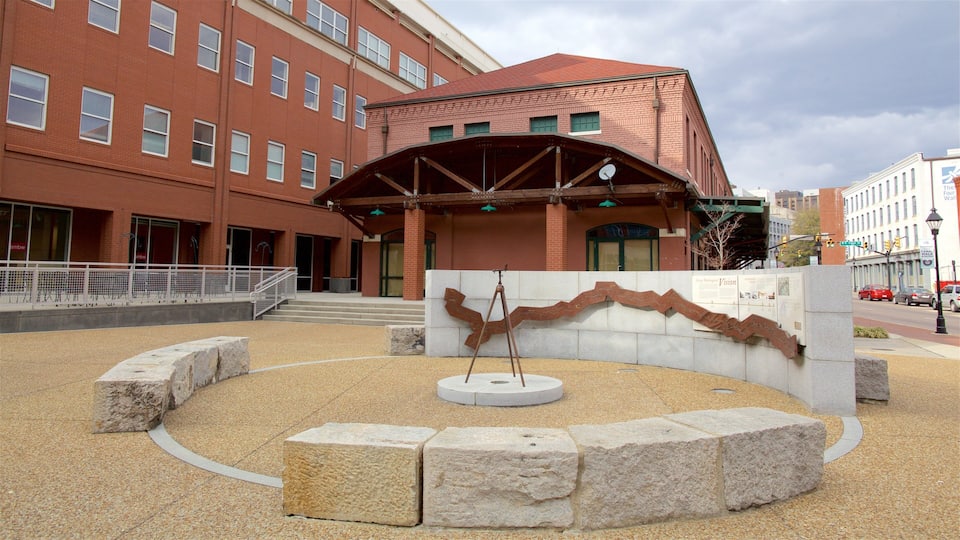 A plaza with a stone sculpture, a red brick building, and a street with cars.