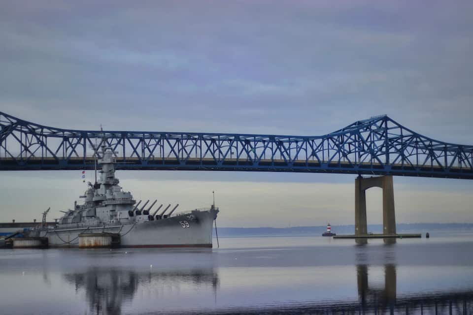 A naval ship docked under a large blue bridge over calm water.