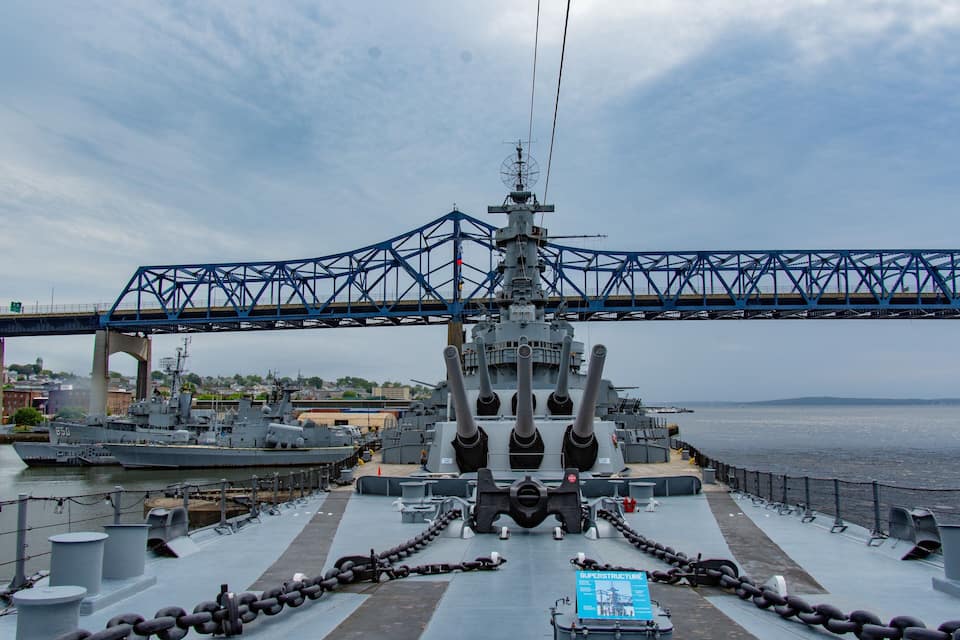 A naval ship docked with a bridge overhead, a cityscape in the background, and a body of water.