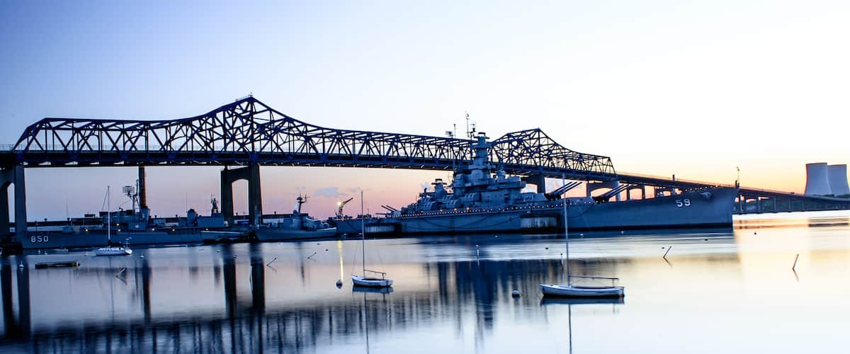 A large naval ship docked under a bridge at sunset.
