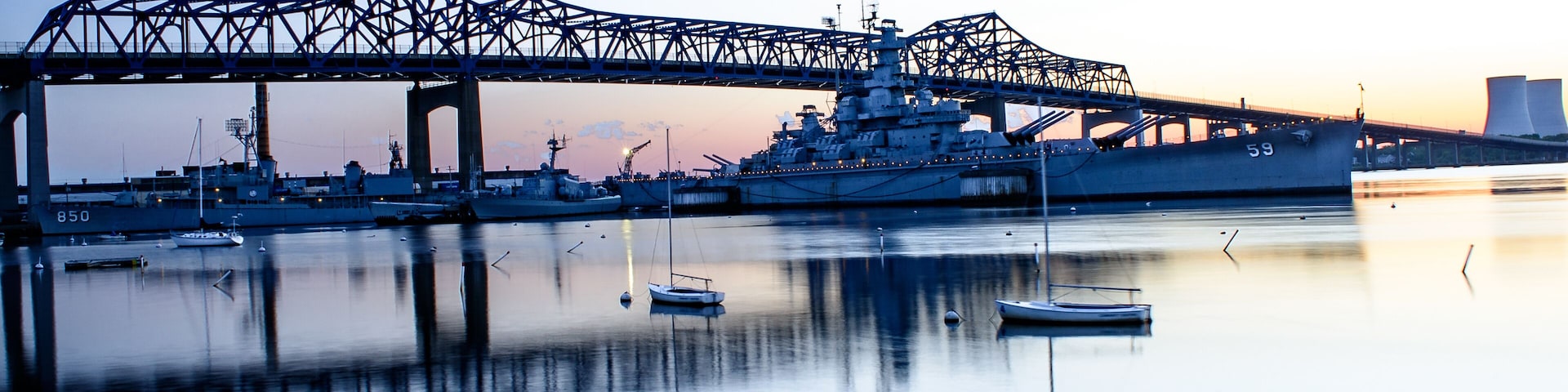 A large naval ship docked under a bridge at sunset.