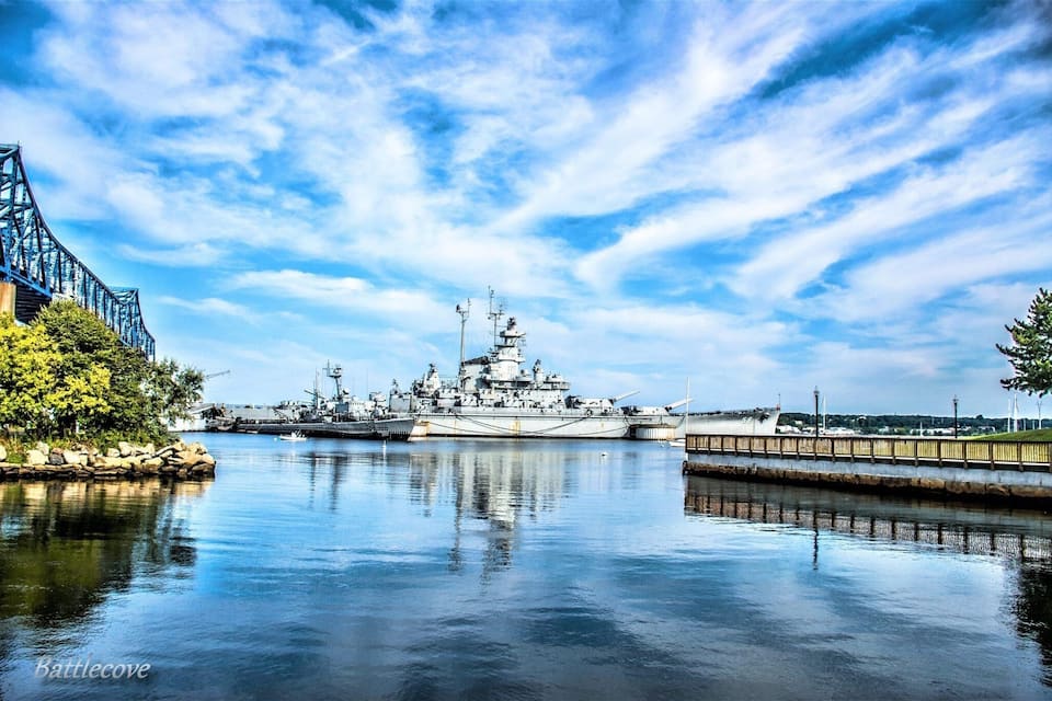 A large naval ship docked at a harbor with a bridge in the background.