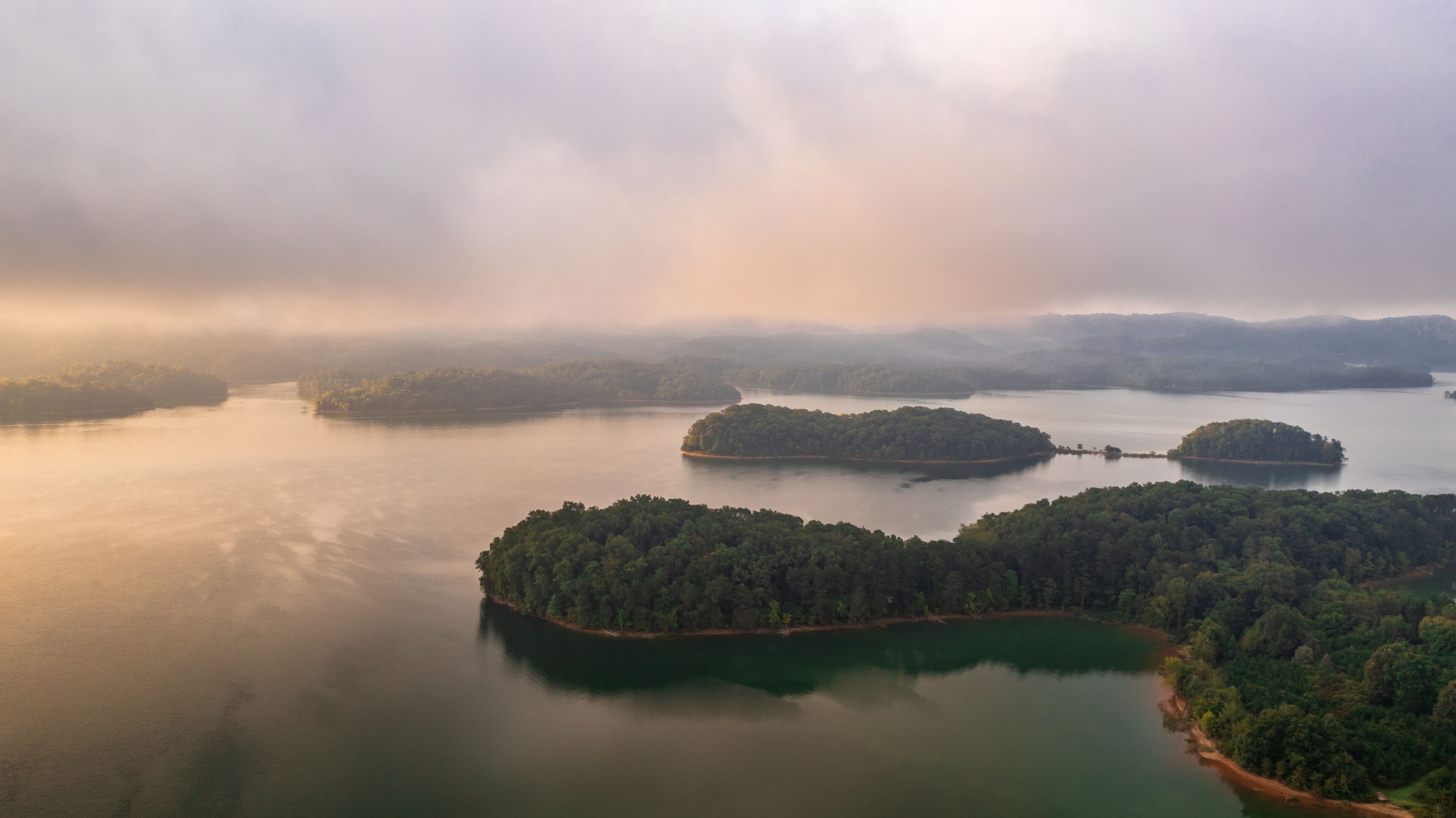 A calm lake surrounded by forested islands and misty mountains in the distance.