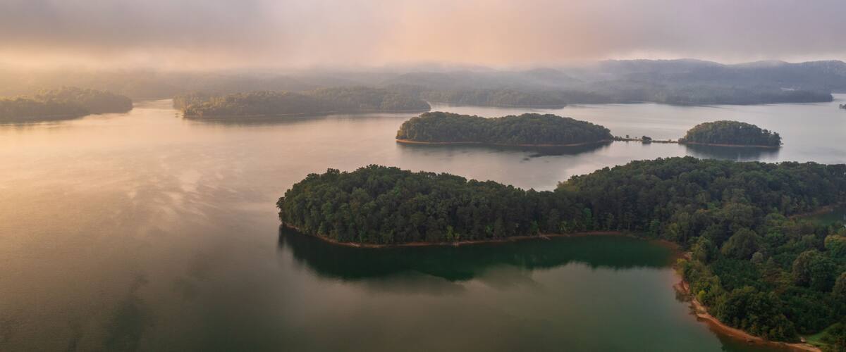 A calm lake surrounded by forested islands and misty mountains in the distance.
