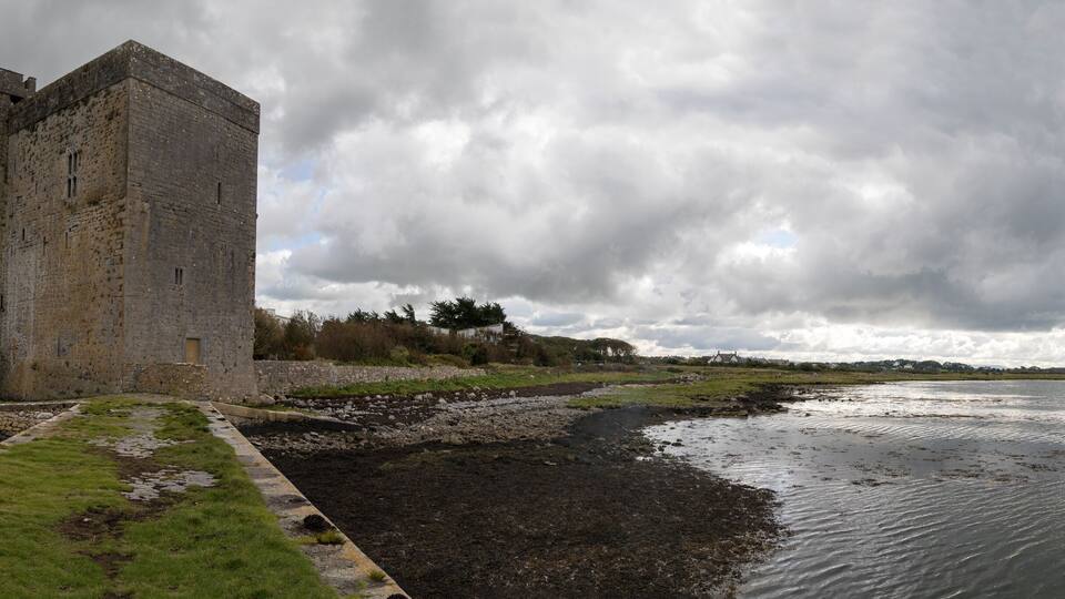 A historic stone tower by a calm river with a cloudy sky.