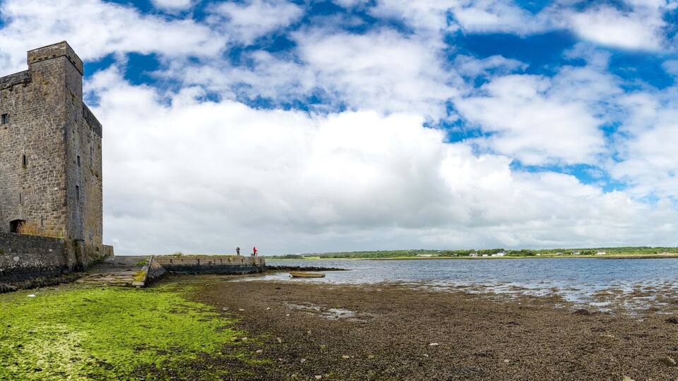 A historic stone tower by a rocky shoreline with a body of water and a cloudy sky.