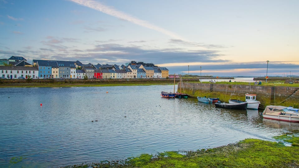 A calm harbor with boats docked, colorful houses along the shore, and a stone wall.