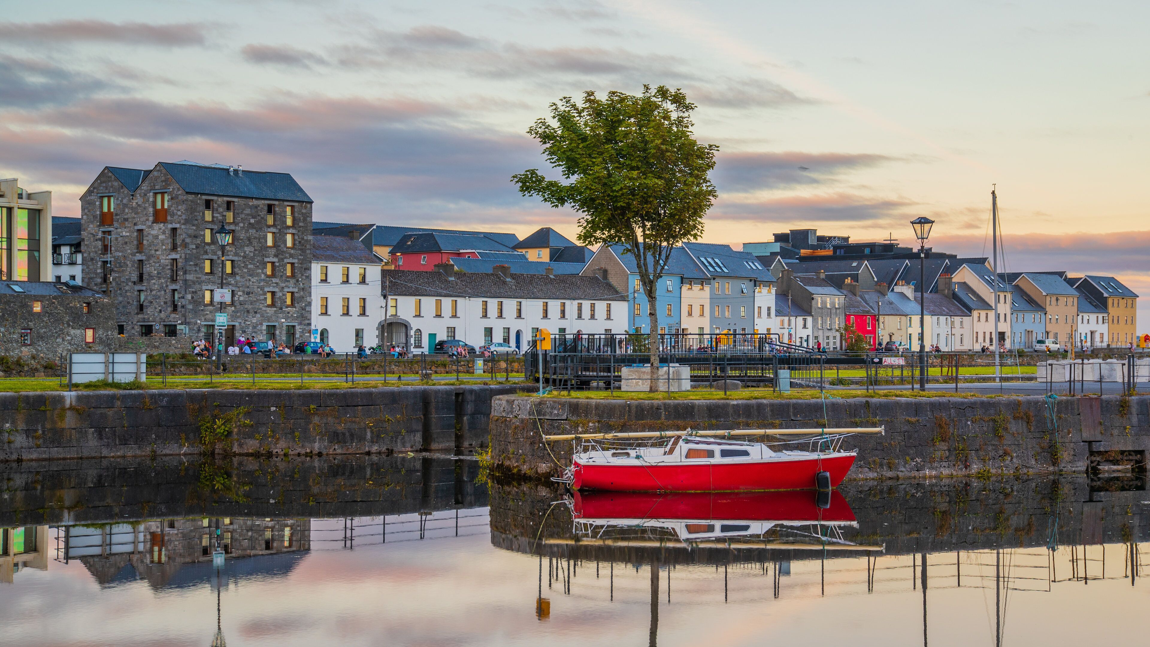 A waterfront with a red boat docked in the foreground, reflecting in the water. In the background, there are colorful buildings and a tree.