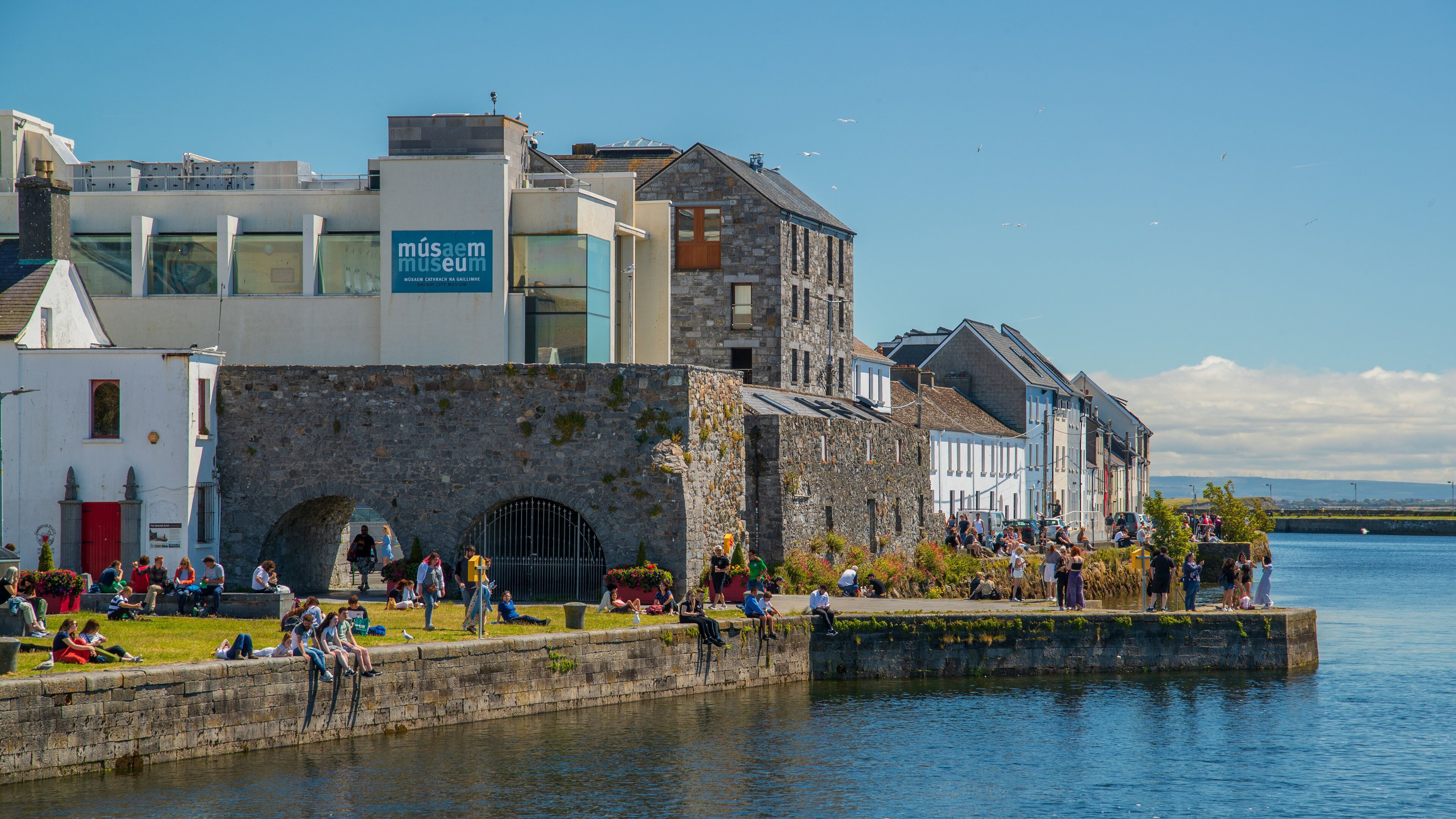 Uferpromenade mit Steinmauer, einem Museumszeichen und Menschen, die am Wasser entspannen.