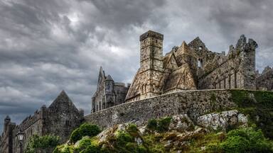 A historic stone castle with multiple towers and a surrounding stone wall, set against a cloudy sky.