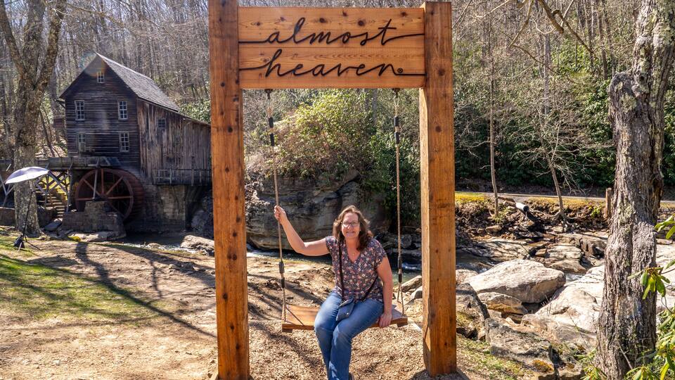 A wooden swing set with a sign that reads "almost heaven" in a wooded area with a rustic building and a water wheel in the background.