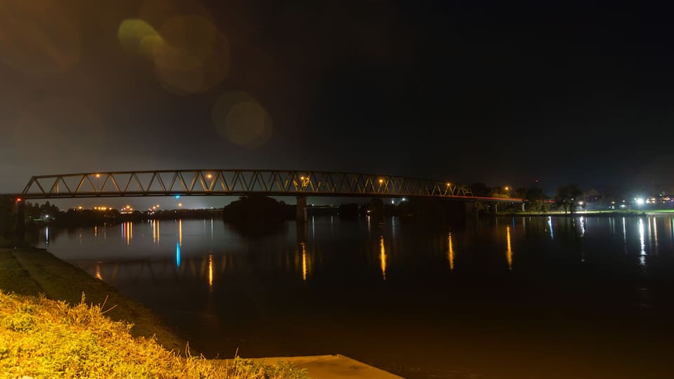 A nighttime view of a bridge over a calm river with city lights reflecting on the water.