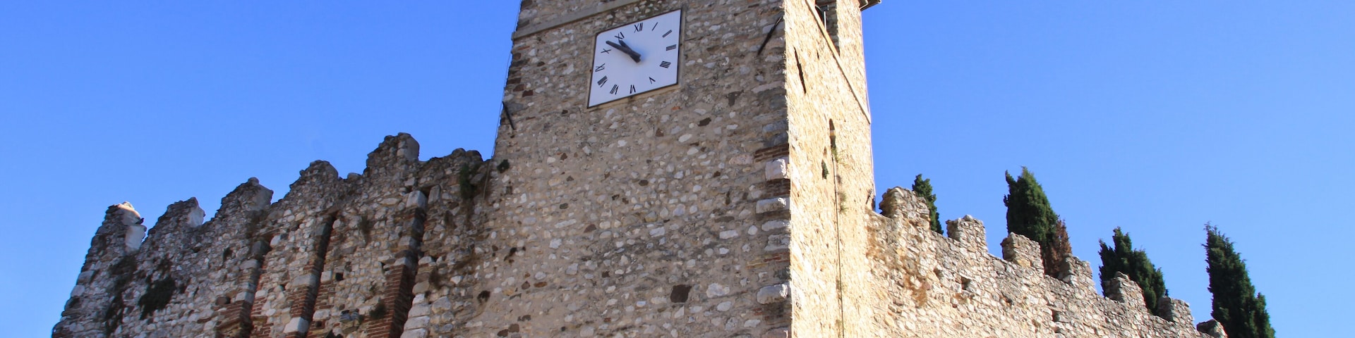 A historic stone tower with a clock, surrounded by greenery and a clear blue sky.