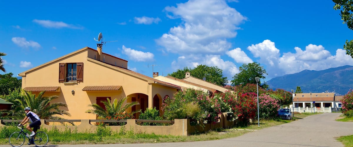 A sunny day in a residential area with a cyclist on the road, a yellow house with red shutters, and mountains in the background.