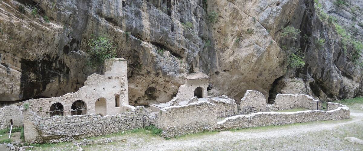 Ruinas antiguas con arcos y un muro de piedra, al pie de un acantilado rocoso.