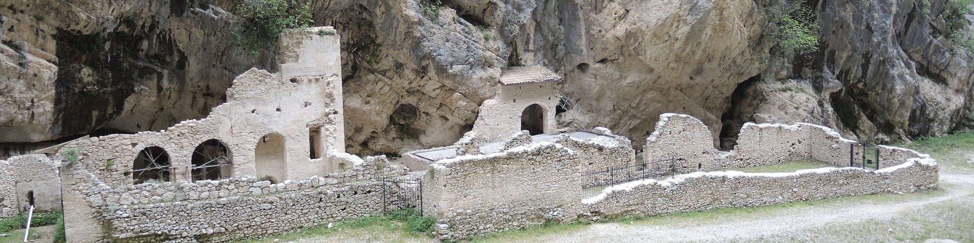 Ancient ruins with arched doorways and a stone wall, set against a rocky cliff.
