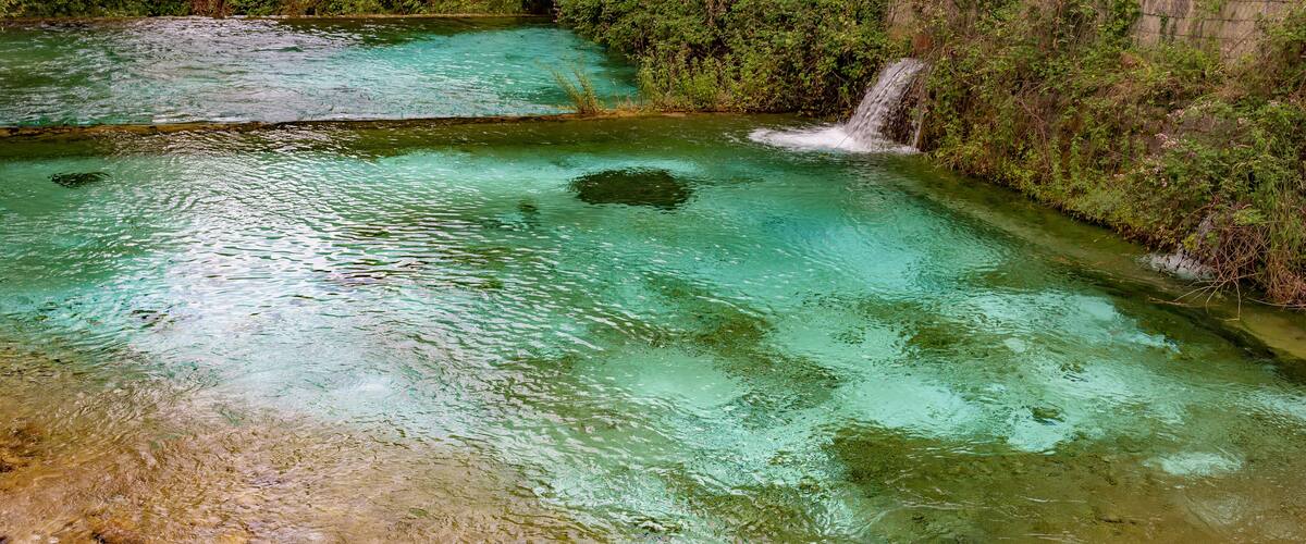 A clear, turquoise pool with a waterfall and lush greenery.