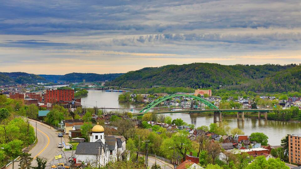 A river with a green bridge, a town with buildings, and green hills in the background.