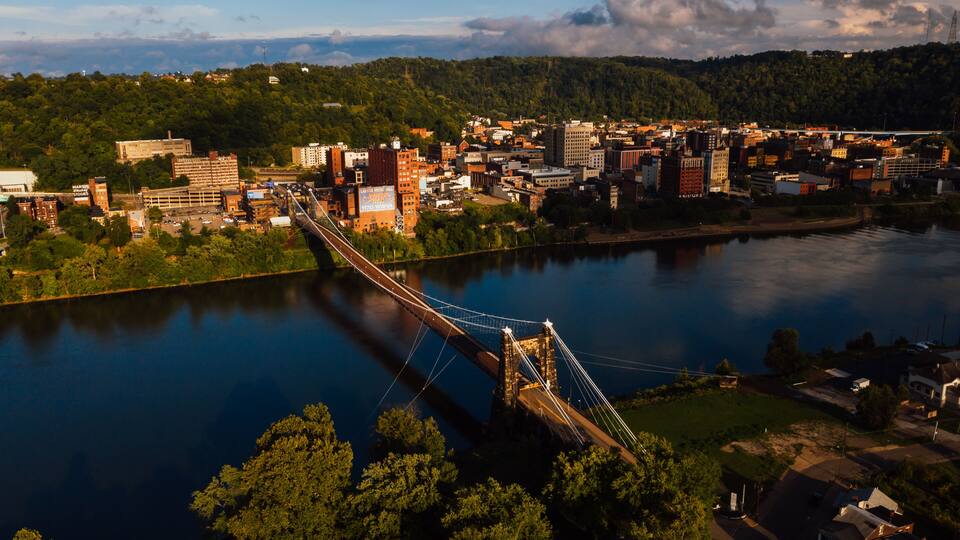 A suspension bridge over a river, surrounded by a cityscape with buildings and a forested hill in the background.
