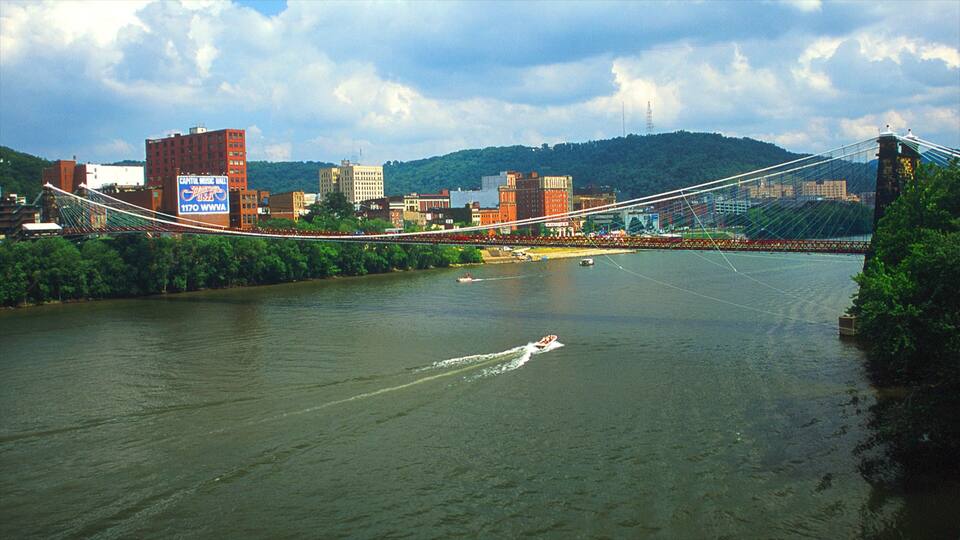 A suspension bridge over a river with boats, surrounded by buildings and hills.