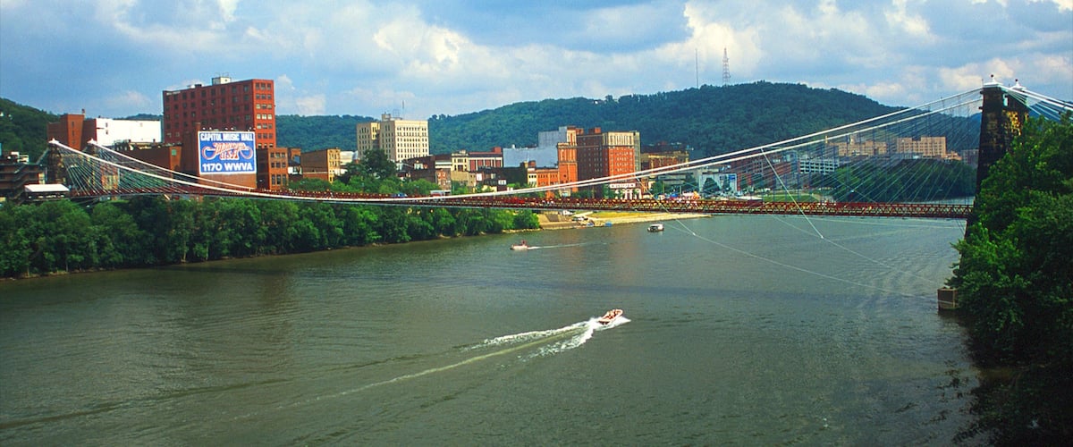 A suspension bridge over a river with boats, surrounded by buildings and hills.