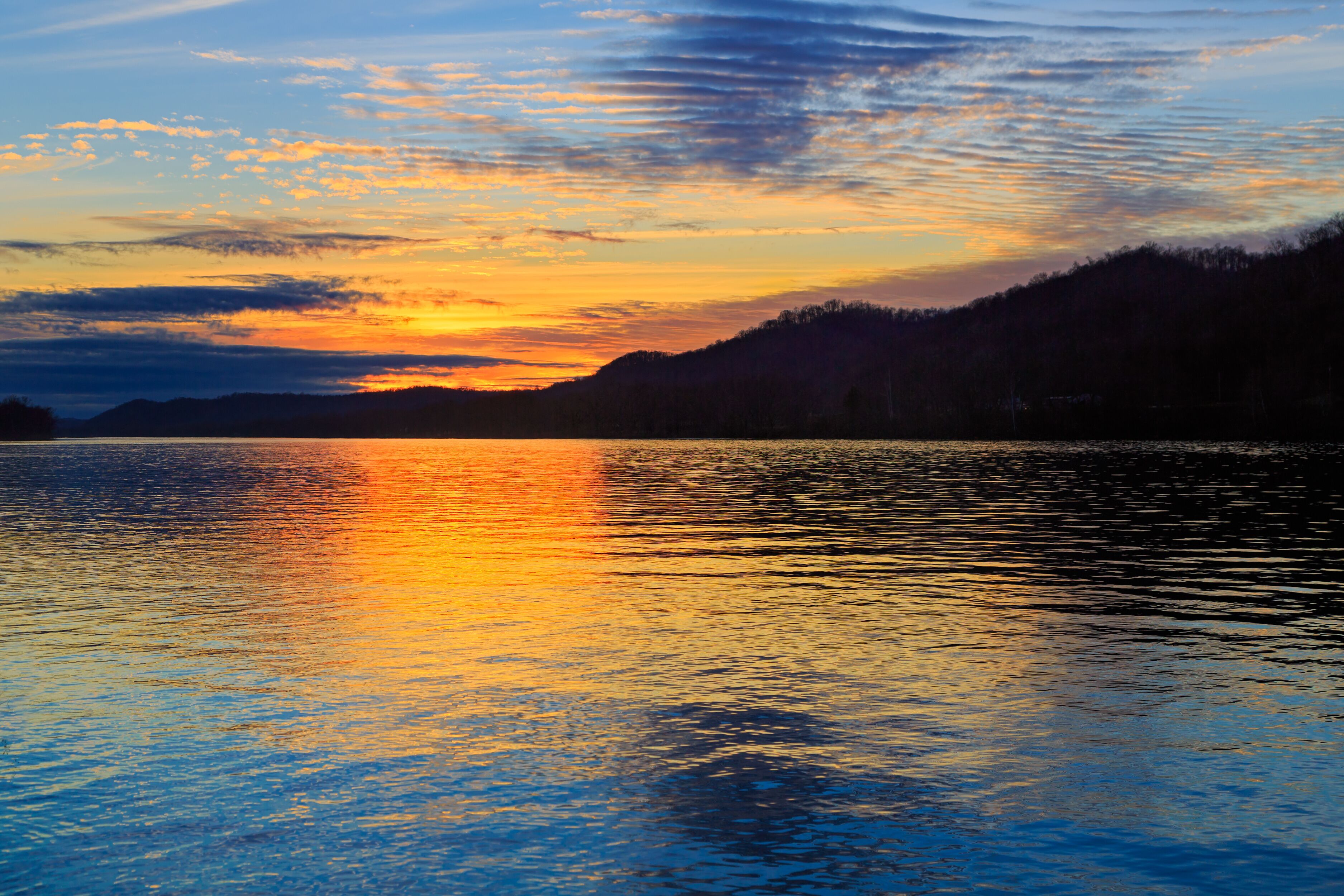 Um pôr do sol sobre um lago calmo, refletindo o céu, cercado por colinas arborizadas.