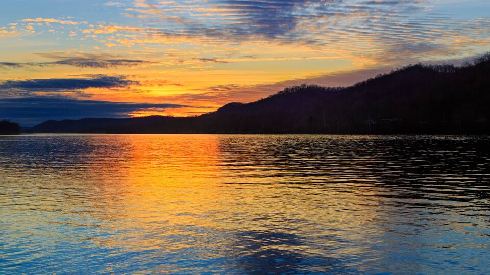 A sunset over a calm lake with reflections of the sky, surrounded by forested hills.