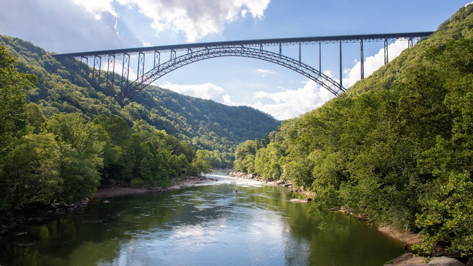 A river with a steel bridge arching over it, surrounded by dense green forest.