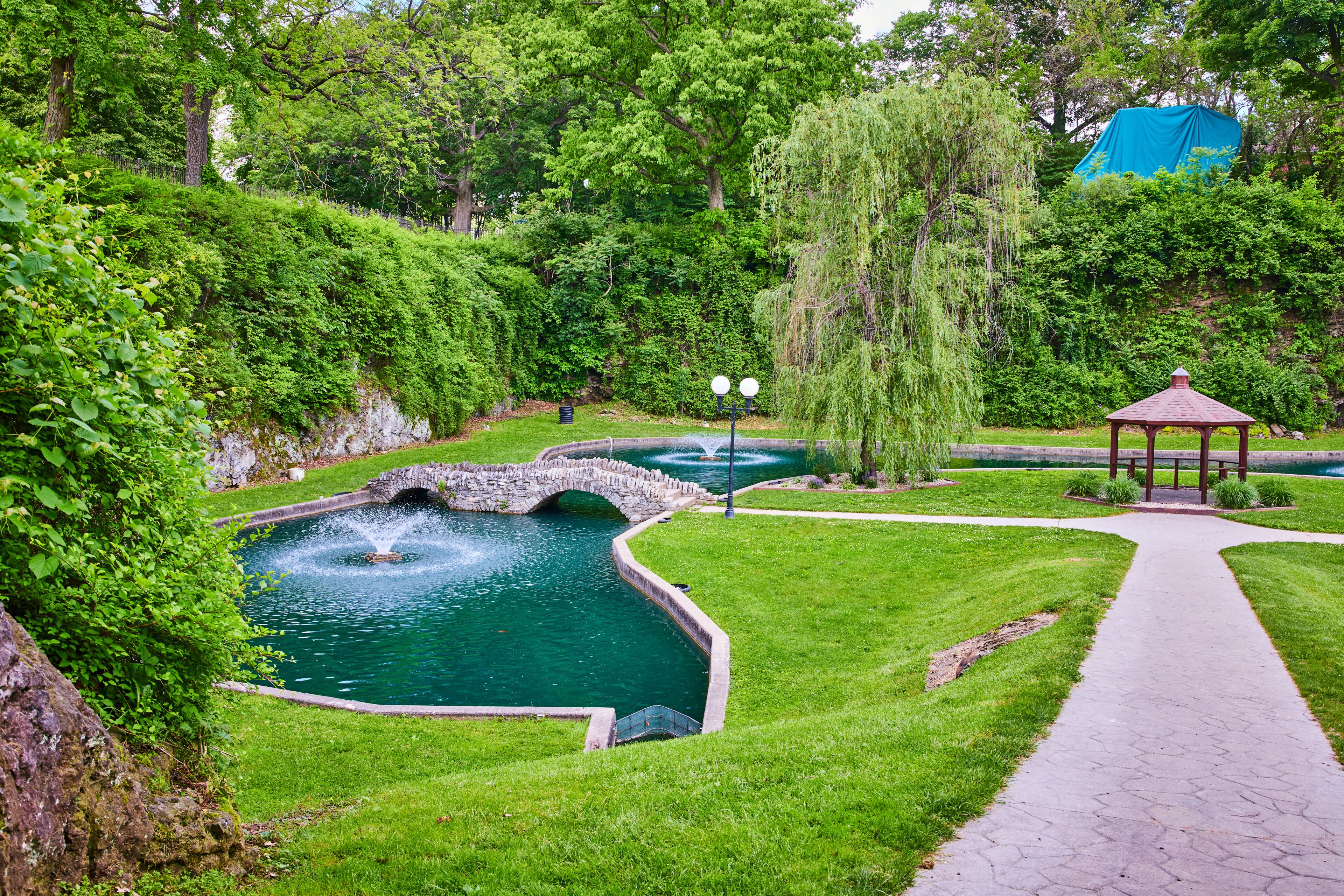 Um jardim paisagístico com lago, uma ponte de pedra, um gazebo e um caminho.