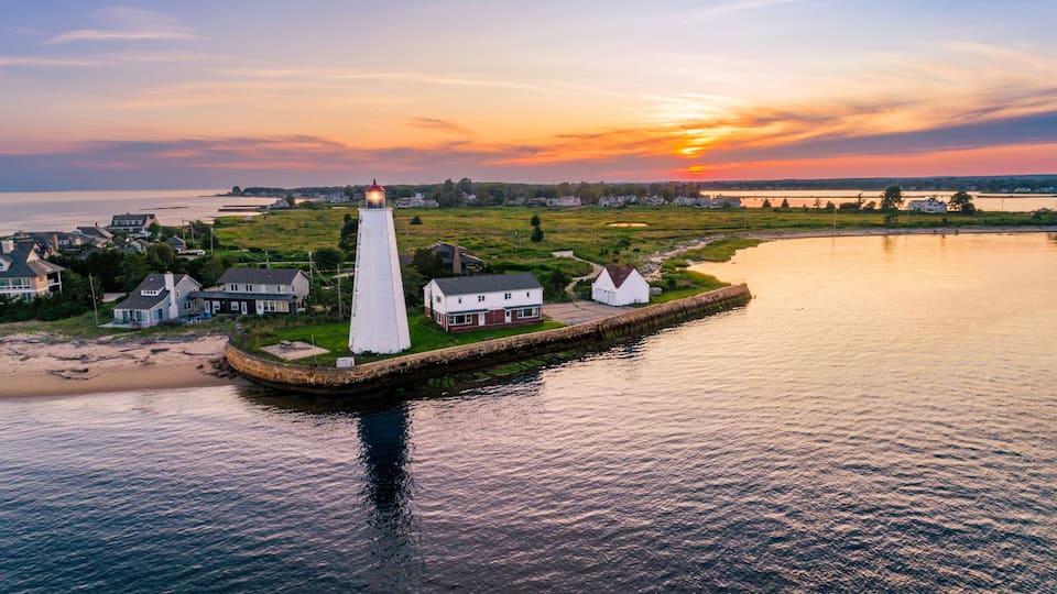 A coastal town with a prominent white lighthouse, houses, and a calm body of water during sunset.